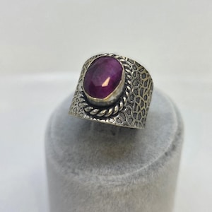 Może przedstawiać: Srebrny pierścionek z dużym, owalnym, ciemnoczerwonym kamieniem szlachetnym. Pierścionek ma teksturowaną obręcz z obramowaniem w kształcie liny wokół kamienia szlachetnego. Obręcz ma wzór plastra miodu.