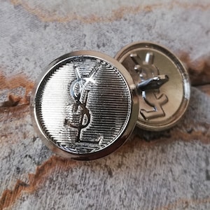 Peut inclure: Deux boutons en métal argenté avec le logo YSL. Le bouton de gauche a un logo YSL en relief et une surface texturée. Le bouton de droite a un logo YSL plat.