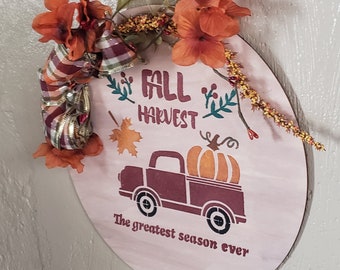 May include: A round wooden sign with the text "Fall Harvest" and a red truck carrying a pumpkin. The sign is decorated with fall-colored flowers and a plaid ribbon. The text below the truck reads "The greatest season ever".