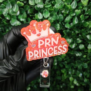May include: A pink glitter badge holder with a white crown and the text "PRN PRINCESS". The badge holder has a clear plastic retractable badge reel with a pink glitter charm.