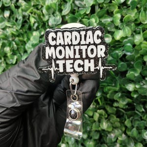 May include: A black and white badge reel with the text "CARDIAC MONITOR TECH" in bold white letters. The badge has a black glitter border and a white background. A silver-colored clip and a black and silver button are attached to the badge.