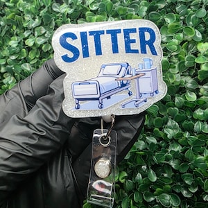 May include: A badge reel with a glittery, light blue and white design. The badge reads "SITTER" in blue letters. The badge features a hospital bed and medical equipment illustration. The reel has a clear plastic strap and a silver button.