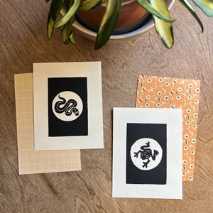 May include: Two greeting cards with black and white linocut prints of a snake and a frog. The cards are on a wooden surface with patterned envelopes and a potted plant in the background. The cards are rectangular with a white border.