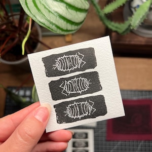 May include: A small, square, white card featuring three black rectangular blocks, each with a white linocut print of a woodlouse. The card is held in a hand, with plants in the background.