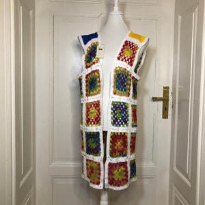 Op de afbeelding: Een lange witte gehaakte vest met een kleurrijk geometrisch patroon. De vest is mouwloos en heeft een V-hals.