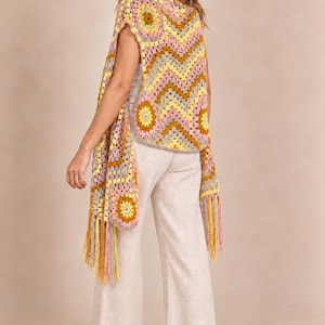 May include: A sleeveless crochet vest featuring a multicolored chevron and circular pattern in shades of yellow, pink, brown, and cream. The garment has a loose, open-front silhouette with long, dangling fringe trim along the bottom hemline.