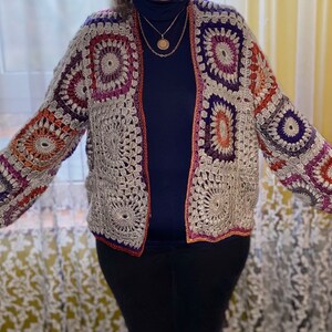 May include: A crocheted cardigan with a multi-coloured geometric pattern. The cardigan is open at the front and has long sleeves. The cardigan is made with a variety of colours, including purple, orange, and grey.