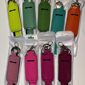 May include: Ten different colored keychain holders for lip balm or other small items. The holders are made of a soft, fuzzy material and have a black strap with a silver key ring.