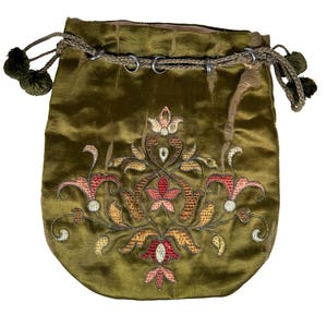 Puede incluir: Una bolsa de terciopelo verde oliva con cordón ajustable y un diseño de bordado floral central en tonos rojo, naranja y crema. La bolsa se cierra con un cordón trenzado que incluye pequeños pompones verde oscuro en los extremos y anillas.
