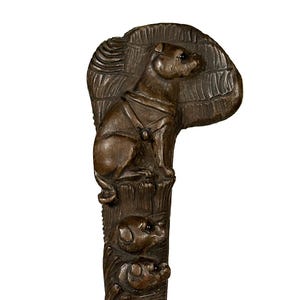 Puede incluir: Un bastón de madera con un perro tallado en la parte superior. El perro lleva un collar y está sentado sobre una base tallada. El bastón es marrón y tiene un acabado liso.