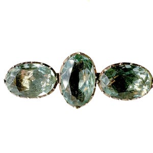 May include: A vintage brooch with three oval-cut gemstones in green and blue hues. The central stone is larger, with two smaller stones either side, set in a silver-toned metal frame. The brooch has a classic, elegant design, suitable for formal wear.