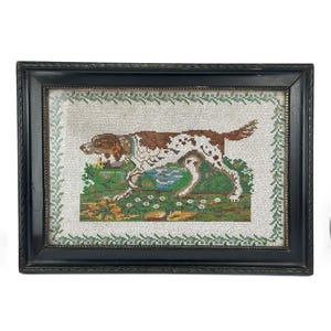 May include: Needlepoint picture of a dog, brown and white, in a field with green grass and a pond. The dog has a blue collar. The black frame has a decorative border. The artwork is framed.