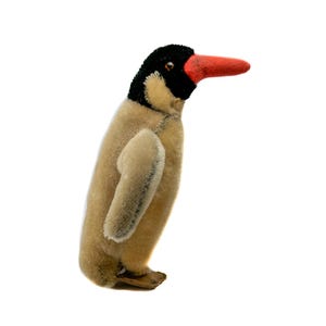 Puede incluir: Un juguete de peluche de pingüino vintage con cabeza negra, cuerpo beige y pico rojo. El pingüino tiene pequeñas alas beige y está de pie. El juguete está hecho de un material suave y afelpado y tiene un aspecto clásico y nostálgico.