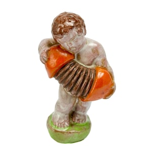 Könnte beinhalten: Eine Keramikfigur eines cherubartigen Wesens mit braunem Haar, das auf einem grünen Sockel steht. Die Figur spielt ein leuchtend orangefarbenes Akkordeon mit dunkelbraunem Balg. Die Skulptur hat eine glänzende Oberfläche und dezente Hauttöne.