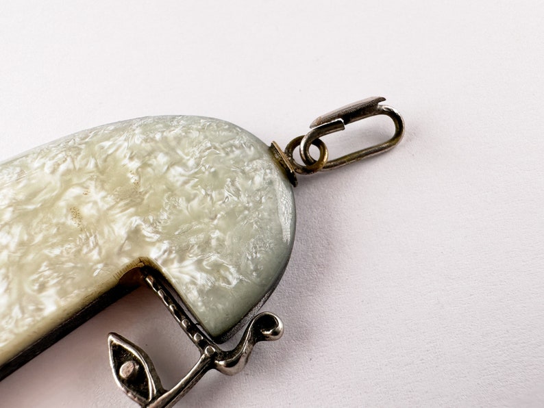 Puede incluir: Un colgante blanco y plateado con un patr&oacute;n de remolino y un cierre plateado. El colgante est&aacute; unido a una cadena plateada.
