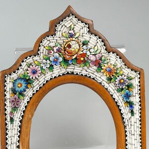 May include: Wooden framed mirror with a mosaic floral design. The frame is a warm brown colour, and the mosaic features a variety of colourful flowers, including blue, pink, and yellow. The mirror has an arched shape.