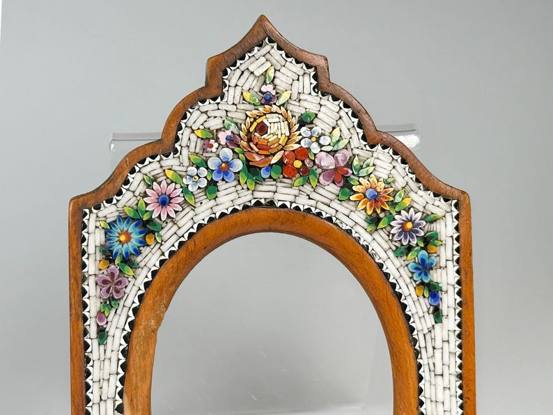 May include: Wooden framed mirror with a mosaic floral design. The frame is a warm brown colour, and the mosaic features a variety of colourful flowers, including blue, pink, and yellow. The mirror has an arched shape.