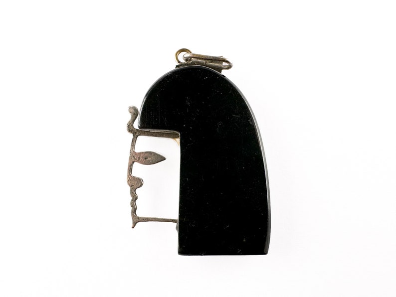 Puede incluir: Un colgante negro y plateado con un dise&ntilde;o de rostro estilizado. El colgante est&aacute; hecho de madera y metal.