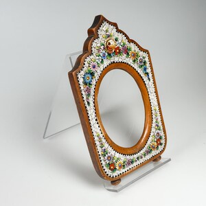 May include: A wooden framed mirror with an oval aperture. The frame is decorated with a mosaic of colourful flowers and white accents. The mirror is displayed on a clear acrylic stand.
