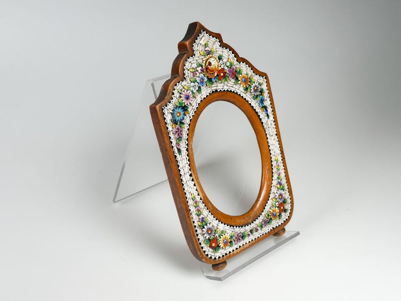 May include: A wooden framed mirror with an oval aperture. The frame is decorated with a mosaic of colourful flowers and white accents. The mirror is displayed on a clear acrylic stand.