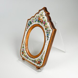 May include: Wooden oval picture frame with a floral mosaic border. The frame is brown with a light wood oval centre. The mosaic features colourful flowers and white accents. The frame is displayed on a clear stand.