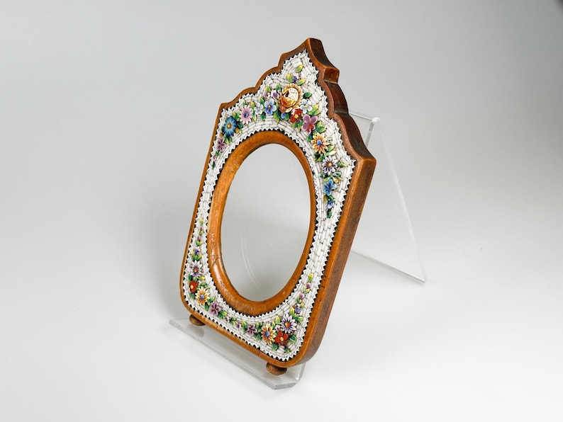 May include: Wooden oval picture frame with a floral mosaic border. The frame is brown with a light wood oval centre. The mosaic features colourful flowers and white accents. The frame is displayed on a clear stand.