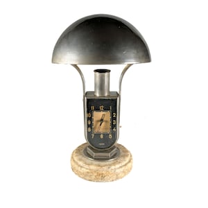 May include: Art Deco desk lamp with a black clock face and a silver dome shade. The clock face displays the numbers 1 to 12. The lamp has a marble base and a silver metal frame. The clock hands are gold.