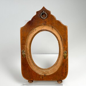 May include: A vintage wooden picture frame with an oval aperture. The frame is a warm brown colour, with a decorative top and small feet. It features a metal ring for hanging and two brass-coloured clasps.