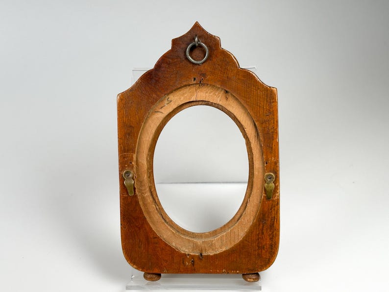 May include: A vintage wooden picture frame with an oval aperture. The frame is a warm brown colour, with a decorative top and small feet. It features a metal ring for hanging and two brass-coloured clasps.