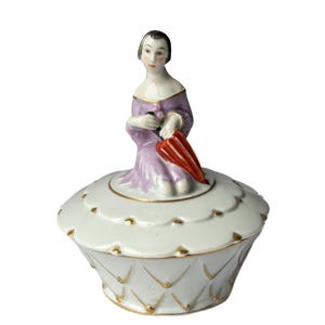 May include: A white porcelain trinket box with a lid, featuring a figurine of a woman in a lavender dress holding a red umbrella. The box has gold accents and a scalloped edge, creating a vintage aesthetic.