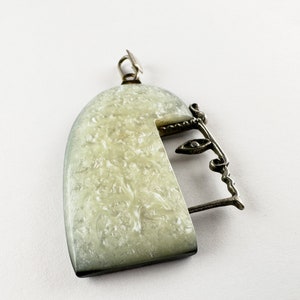 Puede incluir: Un colgante de plata con una forma de semic&iacute;rculo blanco texturizado y un dise&ntilde;o de alambre de plata que se asemeja a una cara.