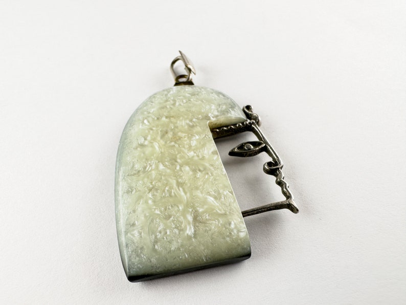 Puede incluir: Un colgante de plata con una forma de semic&iacute;rculo blanco texturizado y un dise&ntilde;o de alambre de plata que se asemeja a una cara.