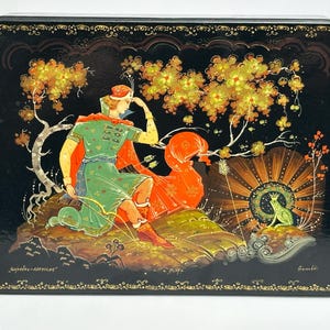 May include: A rectangular black lacquer box featuring a hand-painted scene of a figure in a green tunic and red cape holding a bow, kneeling near a frog wearing a crown. The artwork includes golden trees, Cyrillic text, and ornate gold borders.