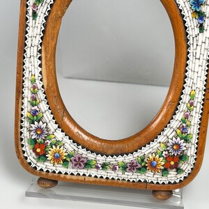 May include: An oval wooden frame with a mosaic border. The mosaic has a white background with colourful floral designs in red, yellow, purple, and blue. The frame has a light brown finish and rests on two small wooden feet.