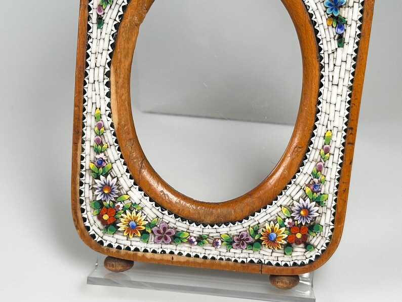May include: An oval wooden frame with a mosaic border. The mosaic has a white background with colourful floral designs in red, yellow, purple, and blue. The frame has a light brown finish and rests on two small wooden feet.