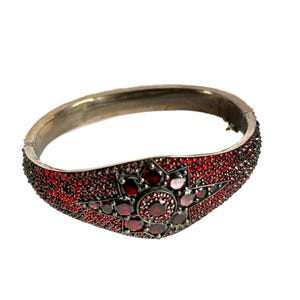 Puede incluir: Brazalete plateado con bisagras y diseño de estrella. El brazalete está incrustado con pequeñas piedras preciosas redondas de color rojo intenso. Un grupo de gemas circulares más grande está en el centro. Una pieza de joyería llamativa.