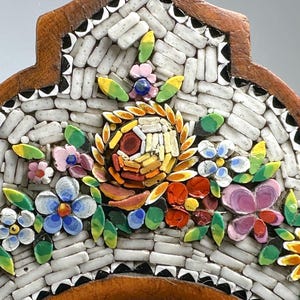 May include: Close-up of a mosaic floral design with a brown wooden frame. The mosaic features white, rectangular tiles and colourful flowers in shades of pink, blue, yellow, and orange. Green leaves accent the flowers.