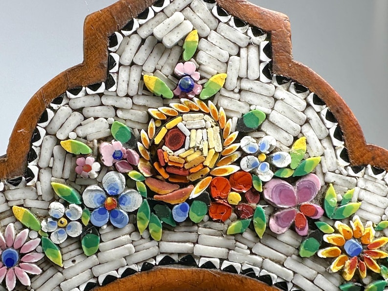 May include: Close-up of a mosaic floral design with a brown wooden frame. The mosaic features white, rectangular tiles and colourful flowers in shades of pink, blue, yellow, and orange. Green leaves accent the flowers.