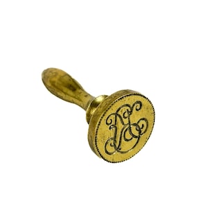 May include: A vintage brass wax seal with a wooden handle. The circular seal features an ornate monogram design. The seal is golden in color and is set against a white background.