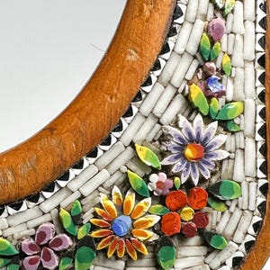 May include: Close-up of a decorative wooden frame with a mosaic floral design. The mosaic features colourful flowers in shades of orange, purple, and red, with green leaves. The frame has a light brown, curved shape.