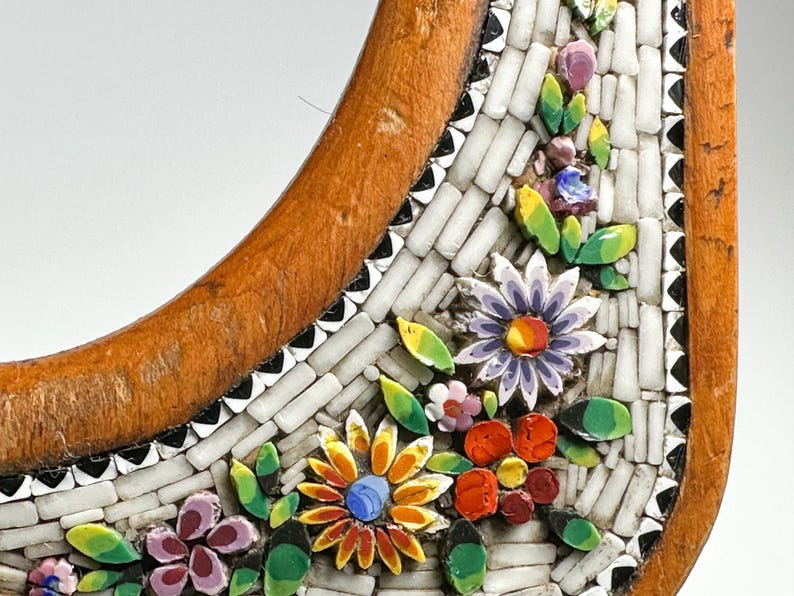 May include: Close-up of a decorative wooden frame with a mosaic floral design. The mosaic features colourful flowers in shades of orange, purple, and red, with green leaves. The frame has a light brown, curved shape.
