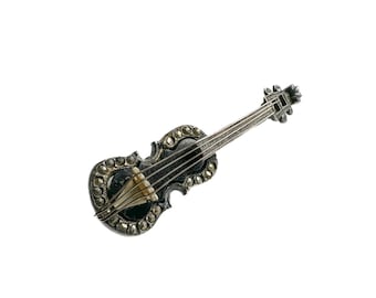 Op de afbeelding: Zilverkleurige vioolbroche met een donkere body en vier zwarte snaren. De broche is versierd met kleine, ronde, heldere stenen langs de randen. Het ontwerp is gedetailleerd en elegant, een klassiek sieraad.