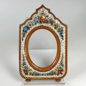 May include: Wooden oval picture frame with a mosaic floral design. The frame has a brown wooden base, a white mosaic border, and colourful flowers. The frame stands on two small wooden feet.