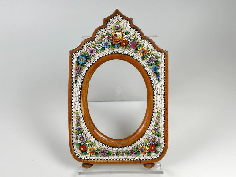 May include: Wooden oval picture frame with a mosaic floral design. The frame has a brown wooden base, a white mosaic border, and colourful flowers. The frame stands on two small wooden feet.