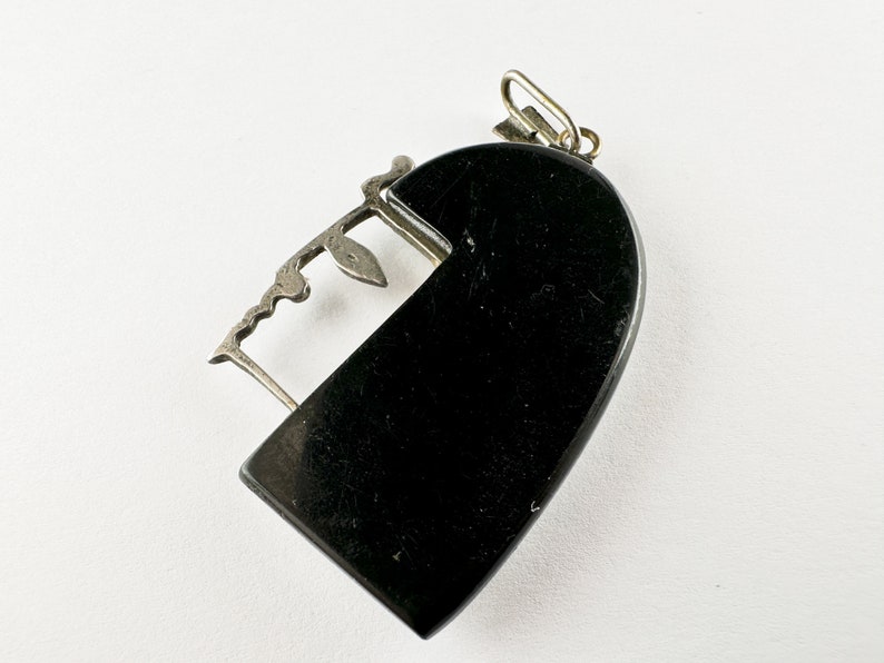 Puede incluir: Un colgante de coraz&oacute;n negro con un dise&ntilde;o de filigrana plateada. El colgante tiene una abertura con bisagra y un peque&ntilde;o bucle en la parte superior para sujetar una cadena.