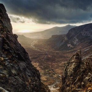 May include: A dramatic view of a valley between two rocky mountain peaks. The valley is covered in brown grass and a stream runs through it. The sky is cloudy and the sun is shining through the clouds.