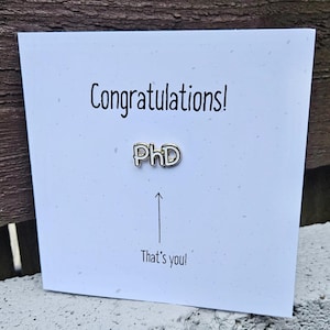 Félicitations ! Carte de remise de diplôme de doctorat avec épinglette en émail « PhD » dorée fixée à la carte. Carte de félicitations pour un docteur en philosophie.