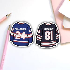 May include: Two sticker decals of hockey jerseys. One is blue with white stripes, the name "HOLLANDER" and the number "24". The other is navy blue with white stripes, the name "ROZANOV" and the number "81".