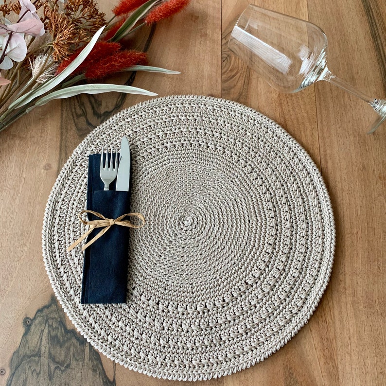 May include: A beige crocheted placemat with a circular design. A black napkin with a silver fork and knife is placed on top of the placemat.