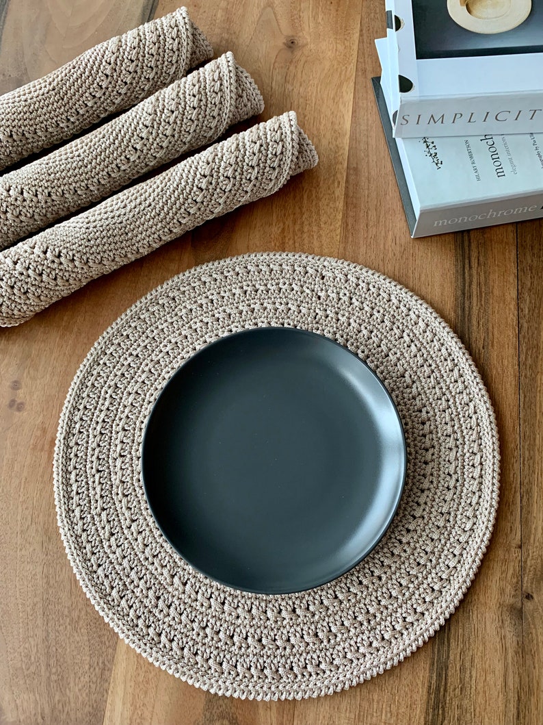 May include: A beige crocheted placemat with a black plate on top. The placemat is round and features a delicate crocheted design. The plate is round and has a smooth surface.